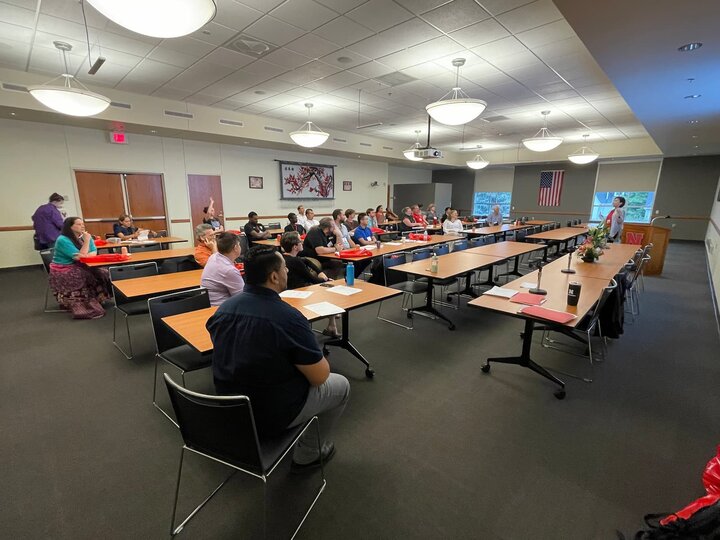 People seated in a conference room with tables, listening to a speaker near a podium.