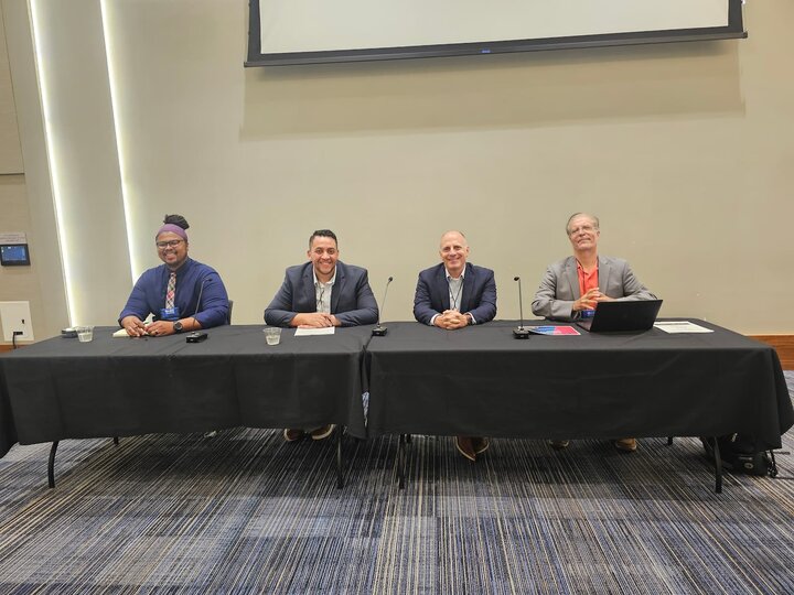 A panel of four men seated behind a table in a conference room with microphones.